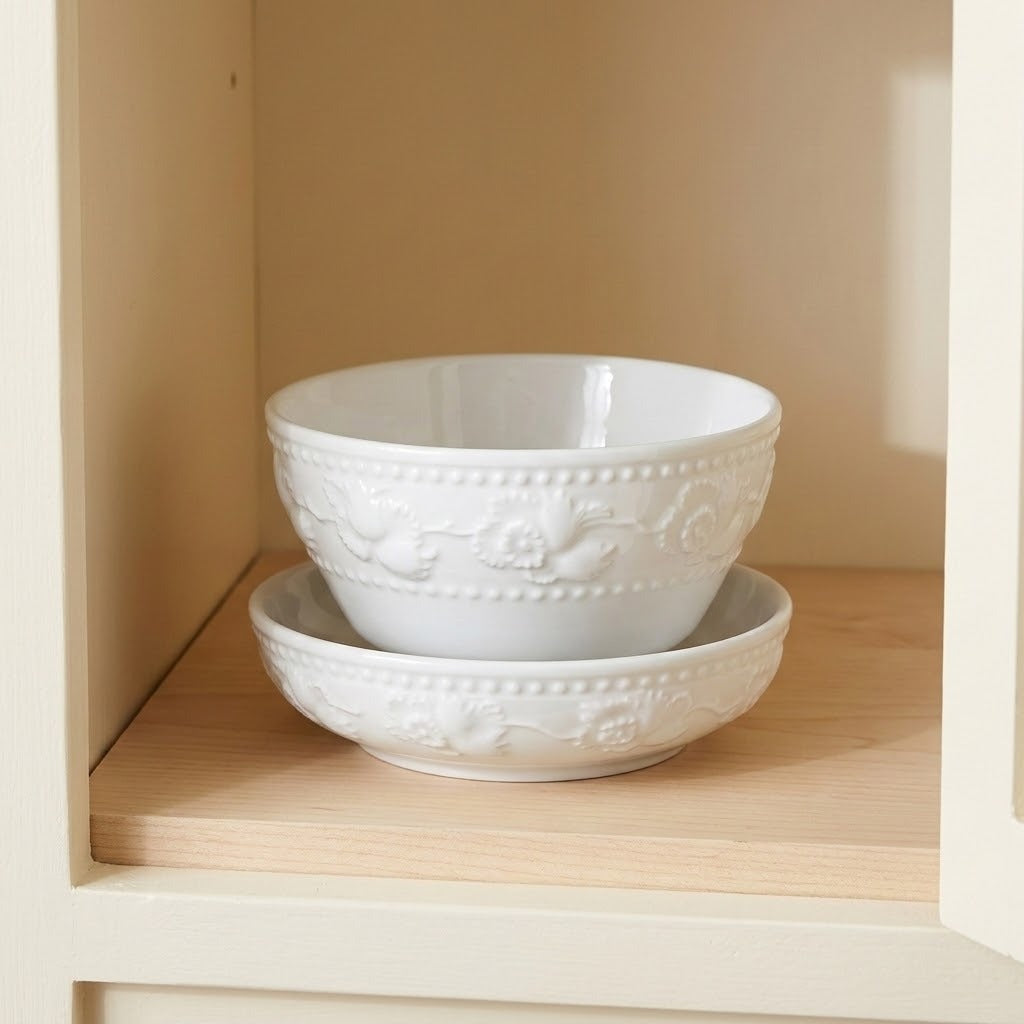 A pair of white ceramic bowls with embossed floral patterns, stacked on a light wood shelf inside a cream-colored cabinet.