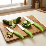 Three green-handled knives with silver blades rest on a wooden cutting board beside sliced cucumber and halved green pepper on a kitchen counter.