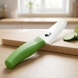 A white-bladed knife with a dimpled green handle rests diagonally across a wooden cutting board beside cucumber slices in a bright kitchen.