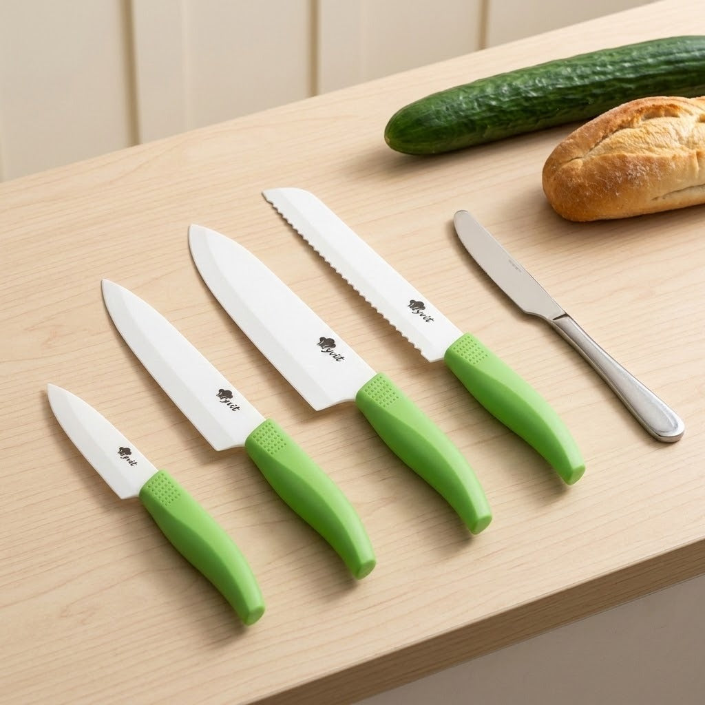 Four white-bladed knives with green handles lie beside a silver table knife, cucumber, and bread roll on a light wooden surface.