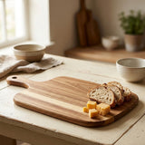 A rectangular wooden cutting board with a handle, shown on a rustic white table in a warm kitchen setting. Slices of bread and cubes of cheese are placed on the board, with bowls and a potted plant blurred in the background.