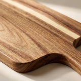 A close-up of a wooden serving board or cutting board with a rich brown and beige wood grain pattern and a curved handle.