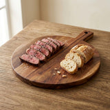 Round brown wooden serving board with a handle holds sliced steak and bread on a rustic wooden table beside a window.