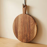 Round brown wooden cutting board hangs by its handle hole from a metal hook against a cream wall above a wooden counter.