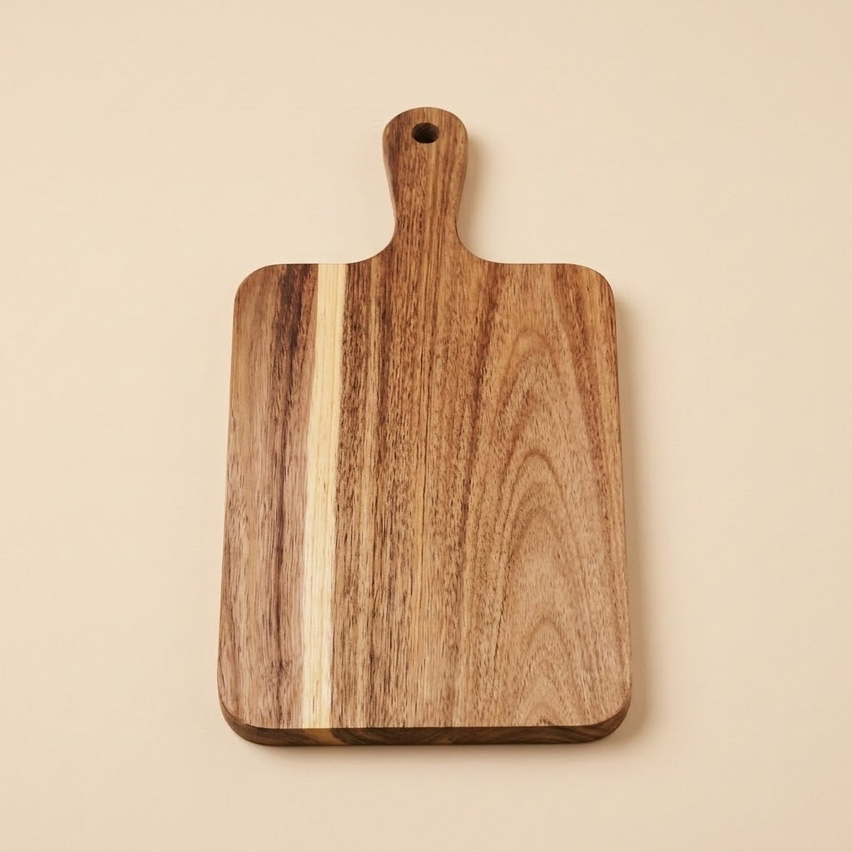 A rectangular wooden cutting board with rounded corners and a handle, featuring natural wood grain patterns in shades of brown and beige, set against a solid light beige background.
