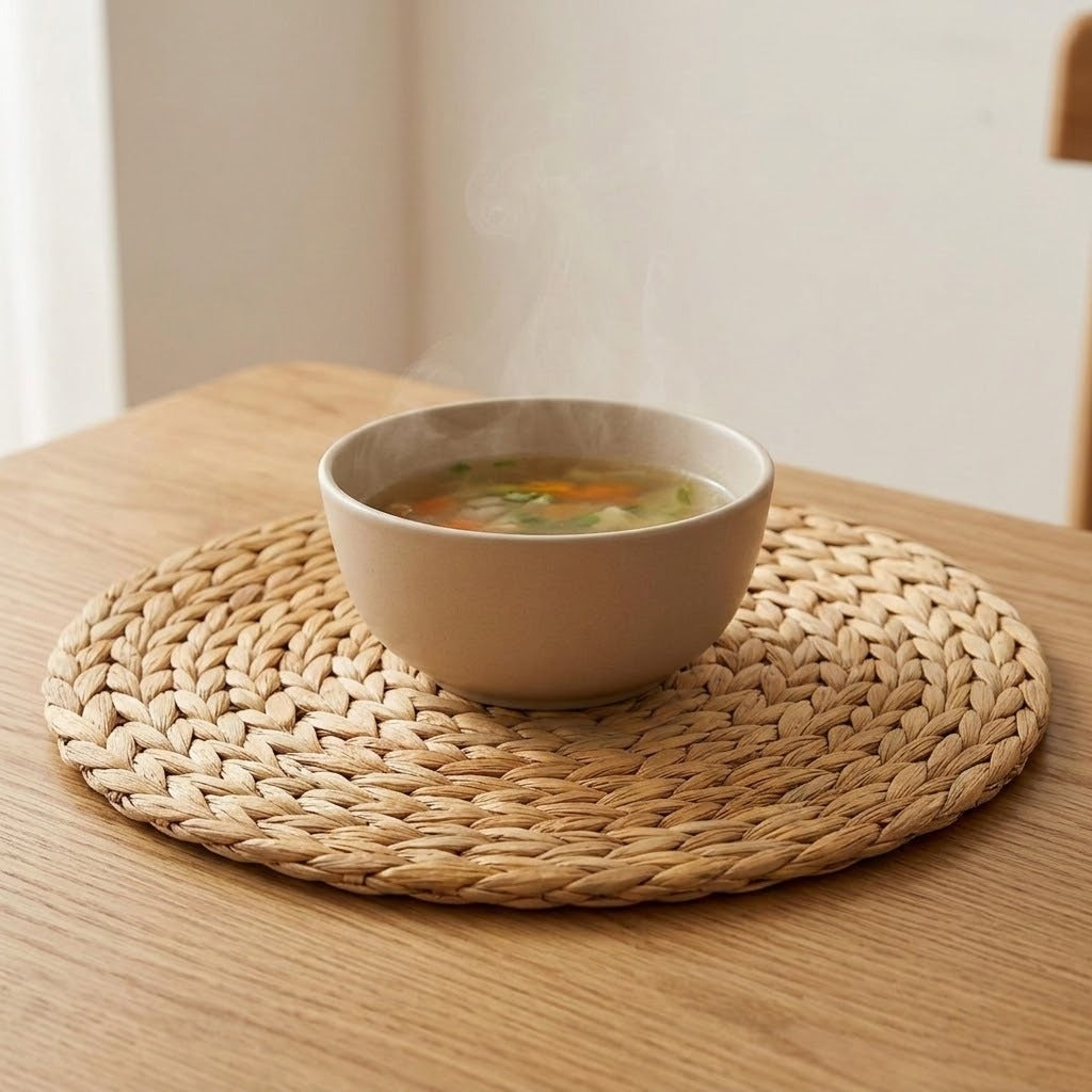 A round tan woven placemat holds a beige bowl of vegetable soup on a wooden table.