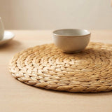 A round tan braided placemat sits beneath a small beige bowl on a light wooden tabletop.