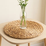 A clear glass vase containing water and green stems stands on a round tan woven placemat atop a pale wooden table.