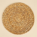 A round tan woven placemat displays concentric braided rings, viewed directly from above against a plain cream background.
