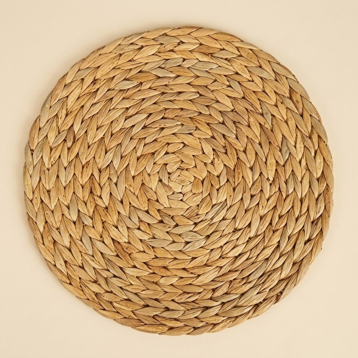 A round tan woven placemat displays concentric braided rings, viewed directly from above against a plain cream background.