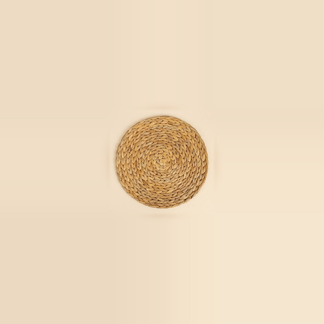 A round woven natural fiber placemat in a light tan color, centered on a solid cream background.