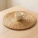 A cream ceramic cup sits in the centre of a round tan woven placemat on a pale wooden table.