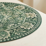 A close-up of a round green placemat with an intricate floral mandala pattern, resting on a light-colored wooden table.