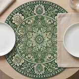 A round green and cream floral placemat lies beneath white plates and a beige napkin on a light wooden table.