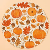 A round cream placemat printed with orange pumpkins, autumn leaves, flowers, and brown acorns lies against a peach background.