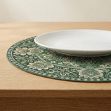 A white plate rests on a round dark green placemat with cream floral patterns on a light wooden table.