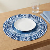 A blue and white geometric placemat holds a white plate beside a water glass and blue napkin on a wooden table.