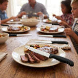 Black-handled serrated knives accompany steak plates on a wooden dining table, with adults and children seated in the background.