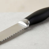 A close-up shows a silver serrated knife blade, shiny metal collar, and curved black handle on a pale surface.