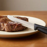 A black-handled knife with a silver serrated blade rests across grilled steak on a cream plate atop a wooden table.