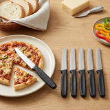 Six black-handled serrated knives sit on and beside a pizza plate, with bread, cheese, and sliced peppers on a wooden table.