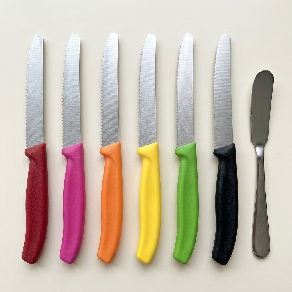 Six silver serrated knives with red, pink, orange, yellow, green, and black handles line up beside a metal spreading knife.