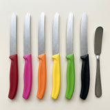 Six silver serrated knives with red, pink, orange, yellow, green, and black handles line up beside a metal spreading knife.
