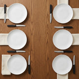 Six white plates with cream napkins and eight black-handled serrated knives are arranged on a wooden dining table.