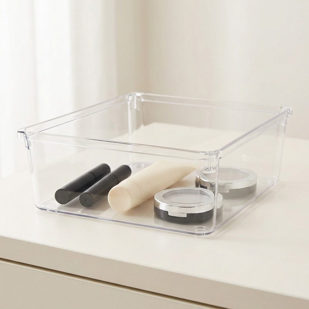 A clear square organizer holds black cosmetic tubes, a cream tube, and round compacts on a white countertop.