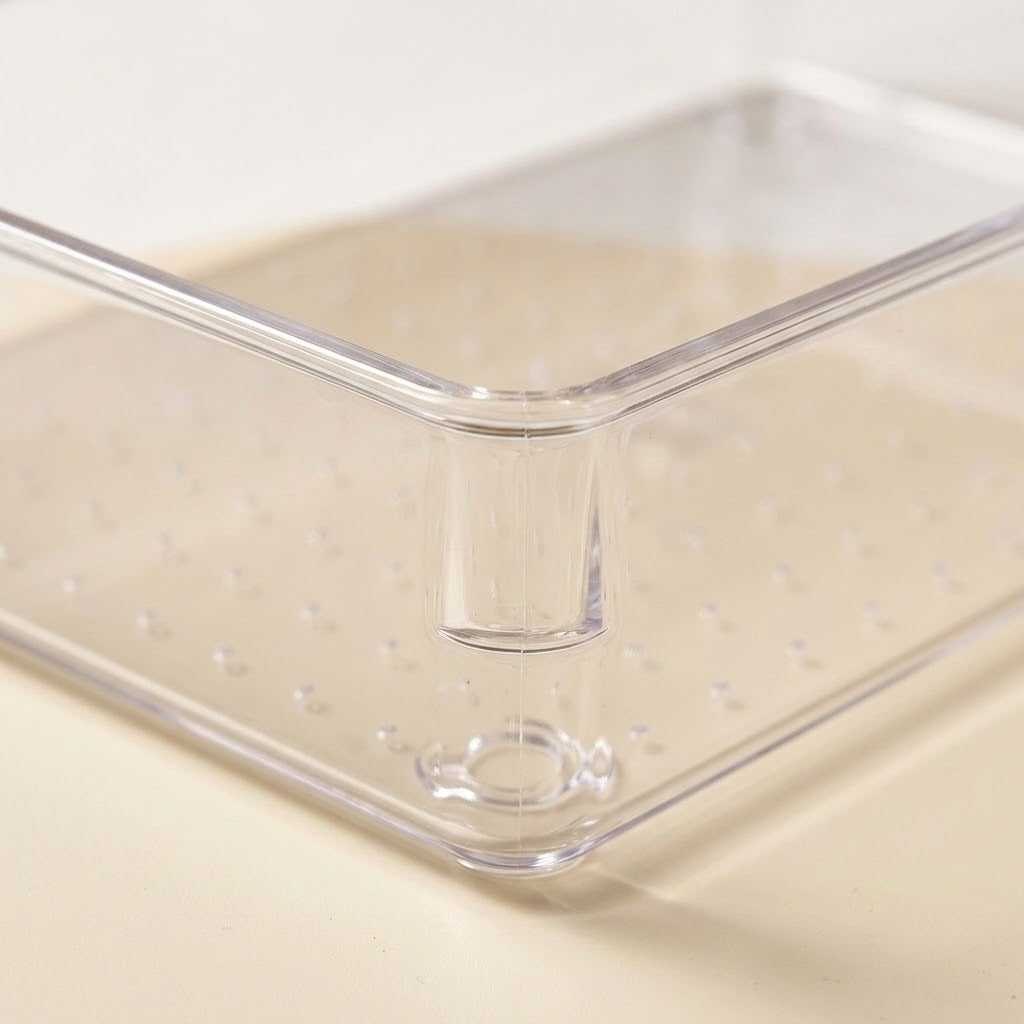 Clear plastic storage container with a raised central divider and perforated bottom, shown on a light beige surface.