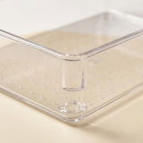 Clear plastic storage container with a raised central divider and perforated bottom, shown on a light beige surface.