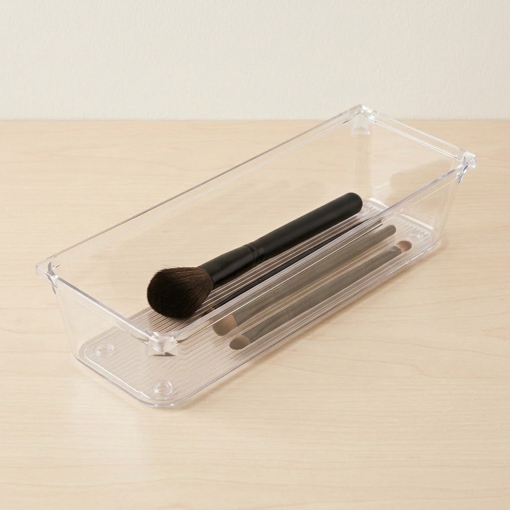 A clear plastic rectangular organizer tray holding several makeup brushes on a light wood tabletop against a neutral wall.