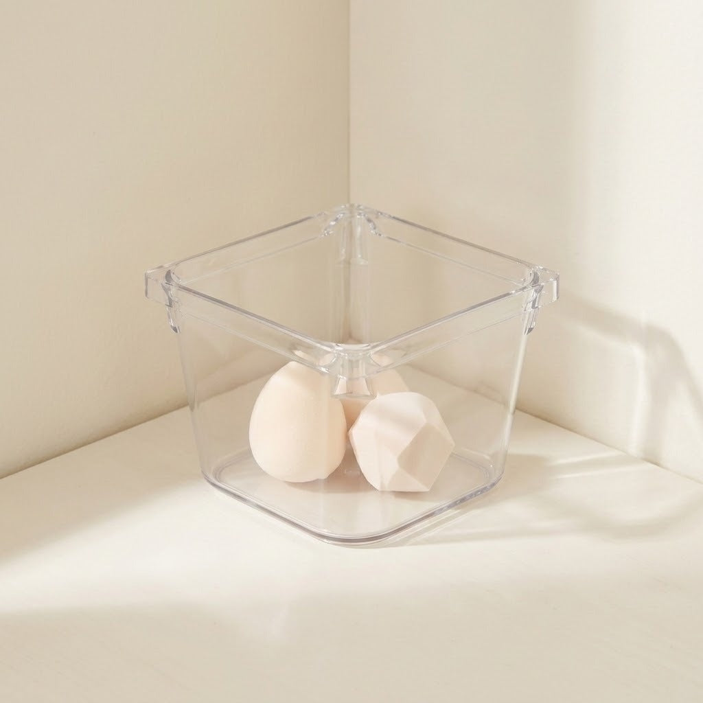 A clear square organizer holds two pale makeup sponges on a white surface against cream walls.