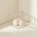 A clear square organizer holds two pale makeup sponges on a white surface against cream walls.