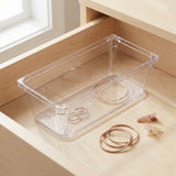 A clear rectangular organizer holds rings and a bracelet inside a light wooden drawer, with gold-toned jewelry beside it.