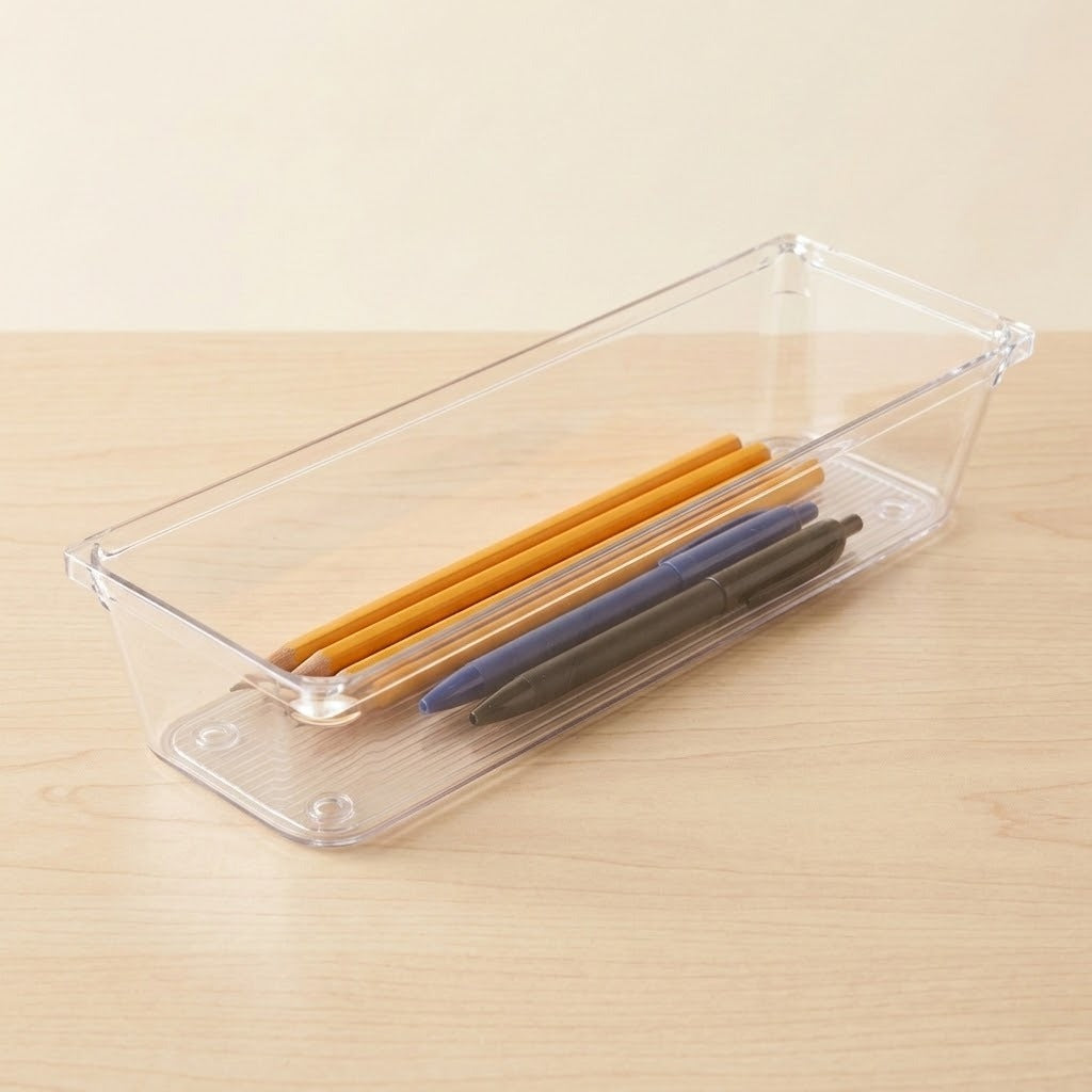 A long clear rectangular organizer holds yellow pencils and blue and black pens on a pale wooden tabletop.
