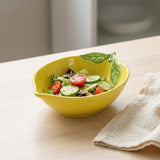 A bright yellow ceramic salad bowl with a leaf-shaped handle and leaf detailing, filled with a fresh salad of tomatoes, cucumbers, and greens, set on a light wood table next to a beige cloth napkin.