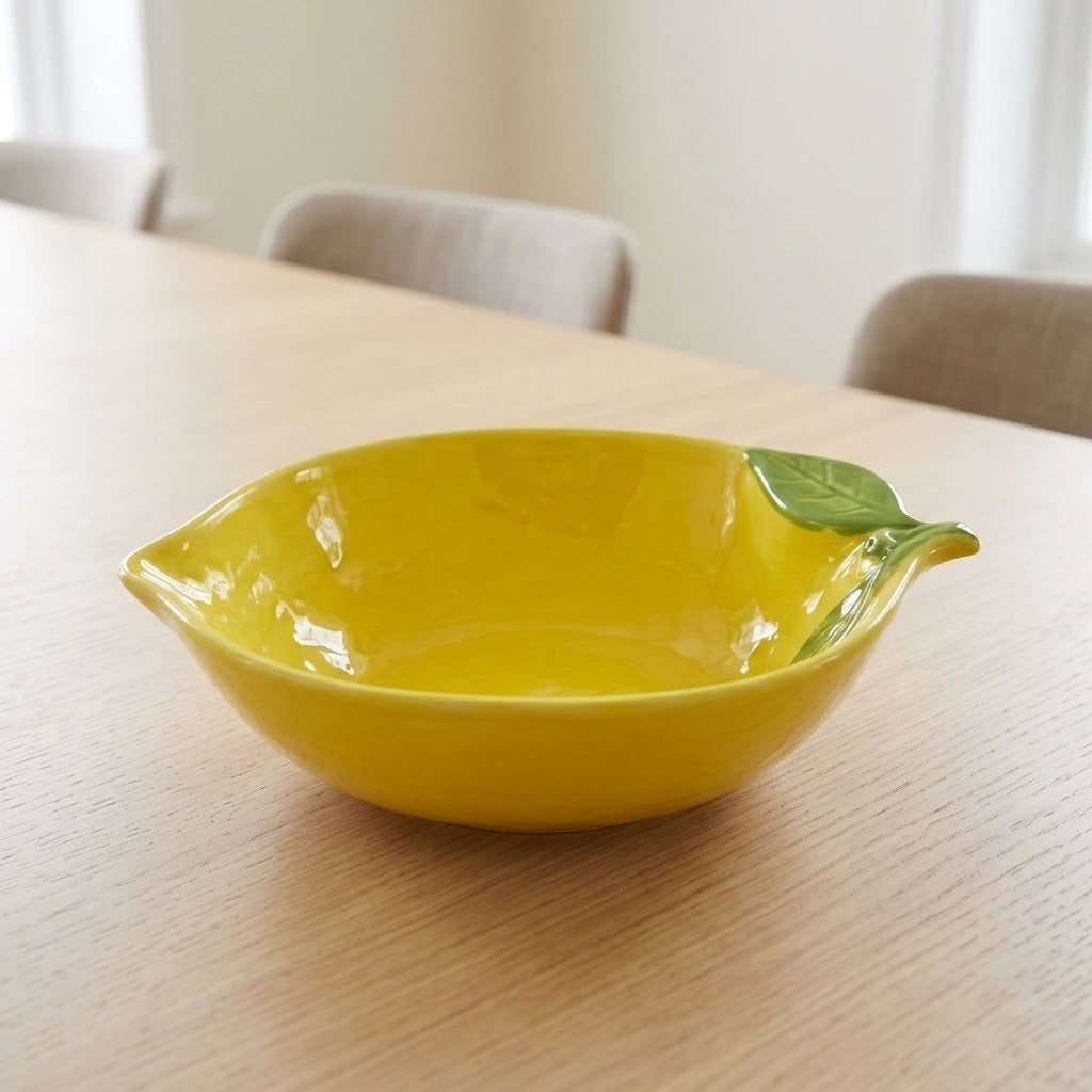 A bright yellow ceramic bowl designed like a lemon, featuring a green leaf accent on the rim, set on a light wood table in a softly blurred dining room setting.