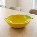 A bright yellow ceramic bowl designed like a lemon, featuring a green leaf accent on the rim, set on a light wood table in a softly blurred dining room setting.