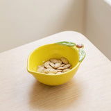 A bright yellow ceramic bowl shaped like a lemon, filled with pumpkin seeds, sitting on a light wood tabletop.