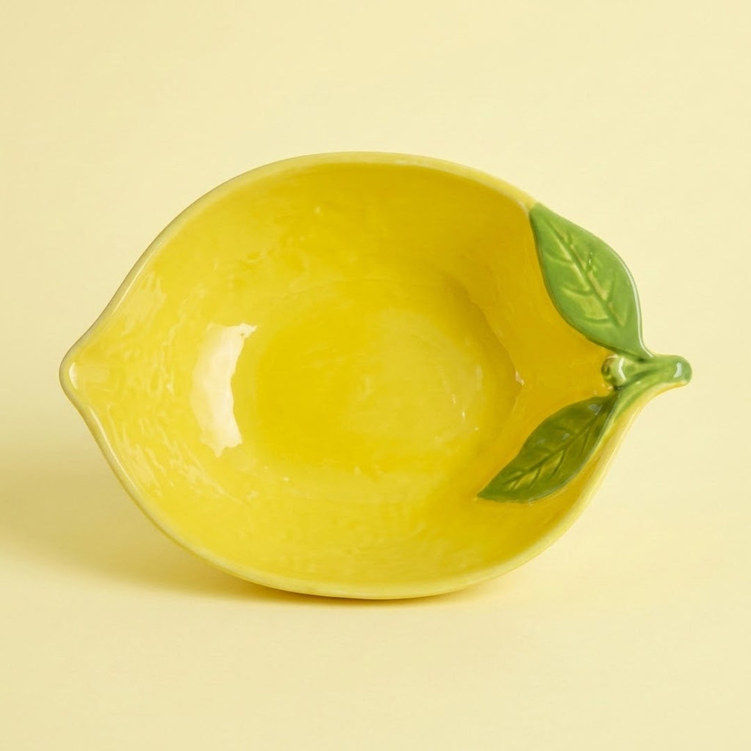 A yellow lemon-shaped ceramic bowl with green leaf and stem details, shown on a solid pale yellow background.