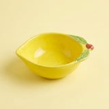 A bright yellow ceramic bowl designed to look like a lemon, featuring green leaf details and a small red stem on the rim, set against a solid pale yellow background.