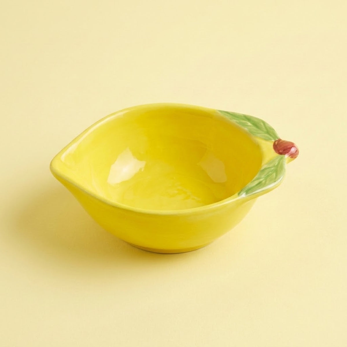 A bright yellow ceramic bowl designed to look like a lemon, featuring green leaf details and a small red stem on the rim, set against a solid pale yellow background.