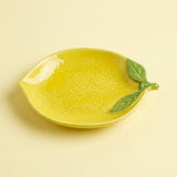 A yellow lemon-shaped ceramic dish with textured detailing and two green leaves on the side, set against a solid pale yellow background.