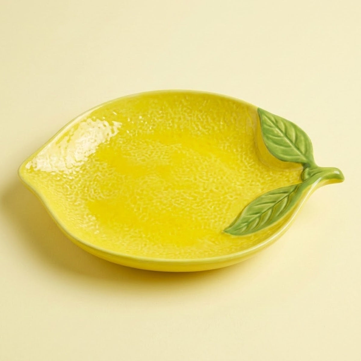 A bright yellow lemon-shaped ceramic dish with textured detailing and green leaf accents on the side, set against a solid pale yellow background.
