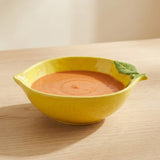 A bright yellow ceramic serving bowl filled with orange soup, placed on a light wood table with a soft beige background. The bowl has small side spouts and a green leaf detail on the right side.