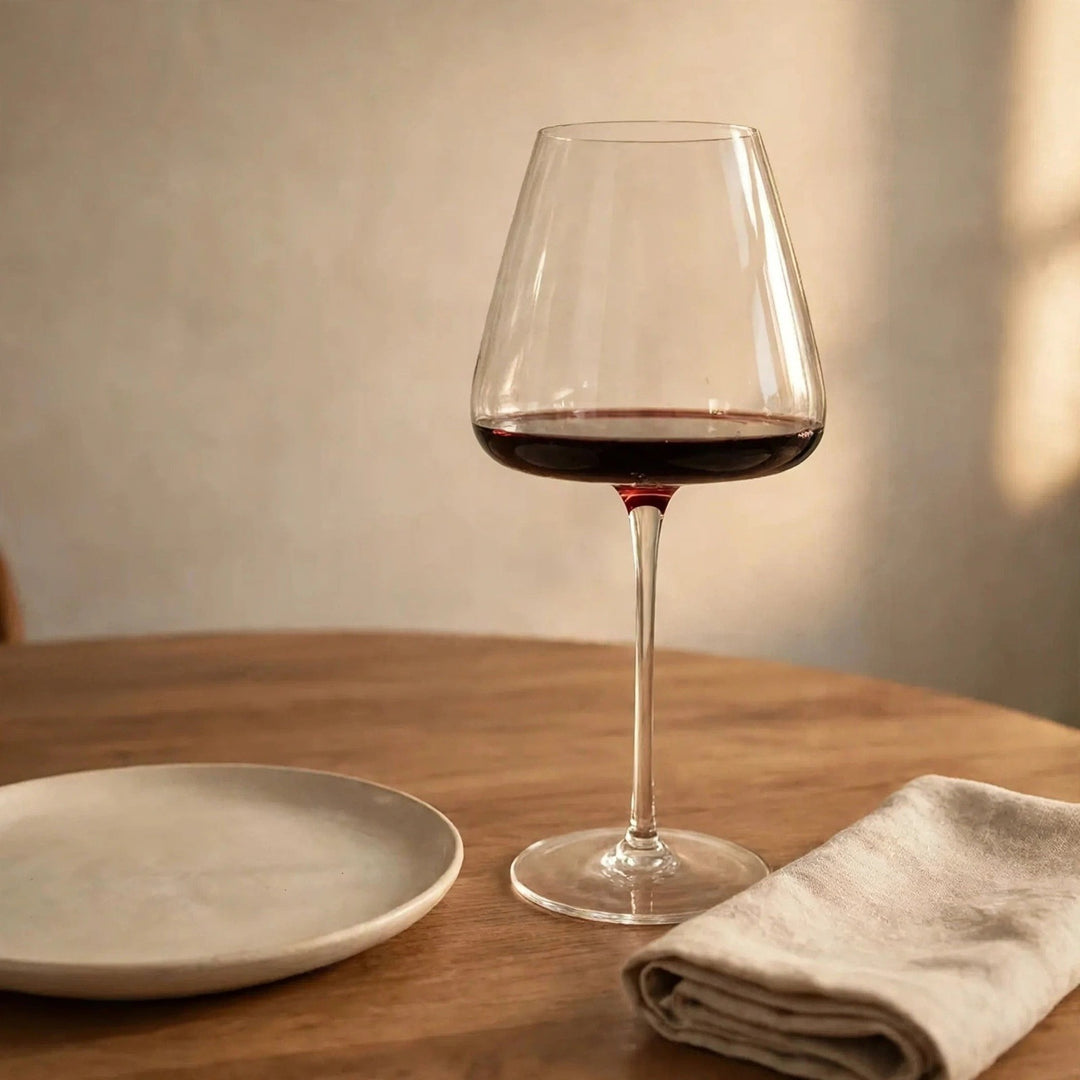 A clear wine glass holding red wine stands on a wooden table beside a cream plate and folded beige napkin.