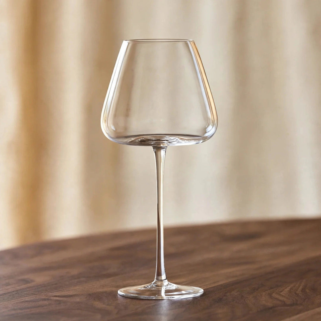 An empty clear wine glass with a tapered bowl and tall stem stands on a dark wooden table against beige curtains.