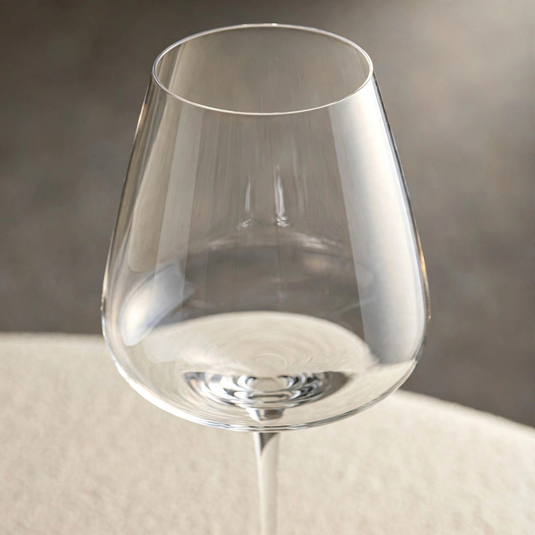 A close-up shows an empty clear wine glass bowl and thin rim against a blurred gray background above a cream surface.