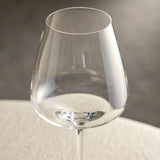 A close-up shows an empty clear wine glass bowl and thin rim against a blurred gray background above a cream surface.